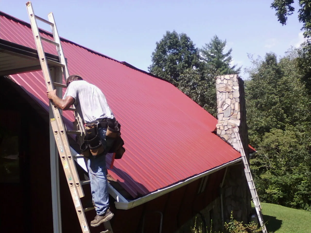 Roofer on a ladder beside a red metal roof and stone chimney — Soffit & Fascia Repair by Roofing Brothers in Mamaroneck